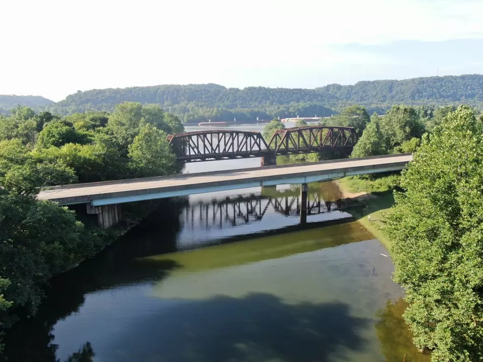 An aerial image taken from a drone showing a bridge over water, with trees on either side and a railroad bridge in the background.